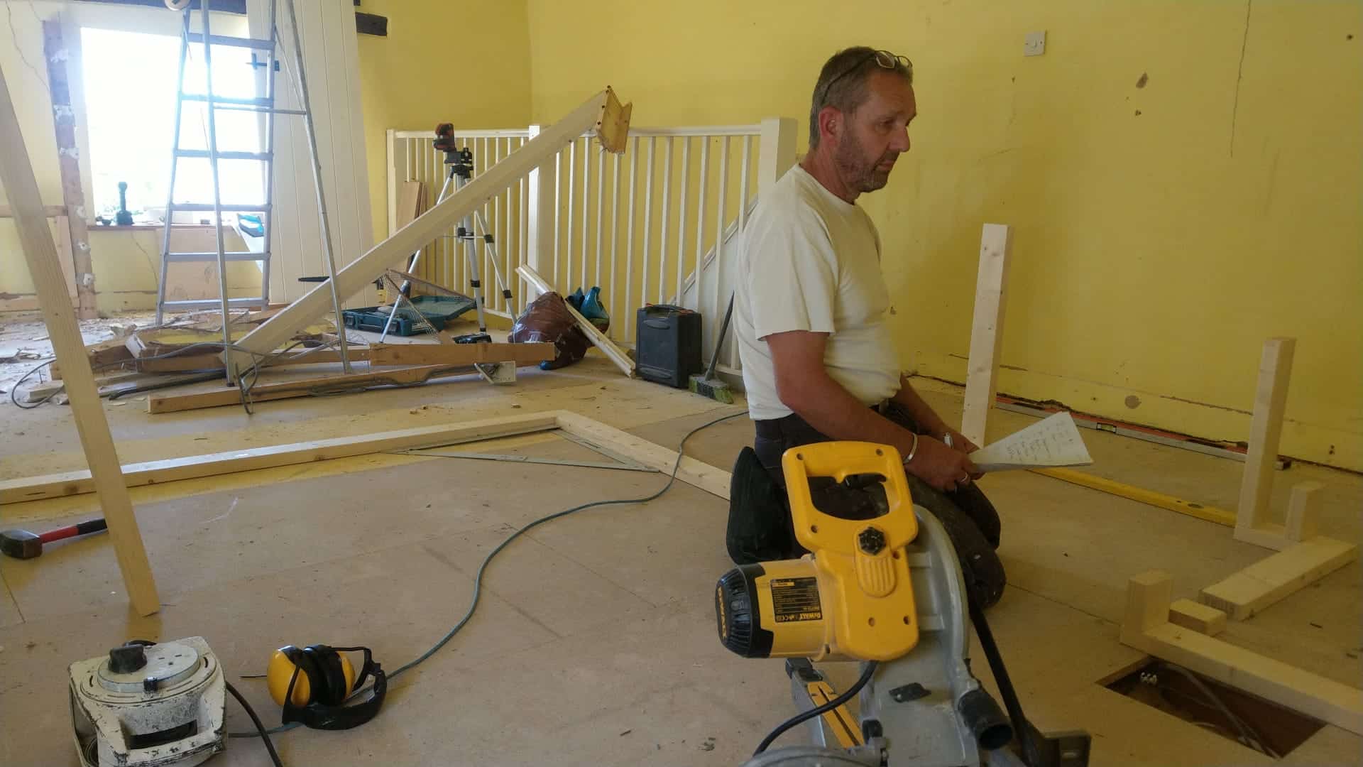 A man works on a property renovation with tools, wood, and a ladder in a room with yellow walls.